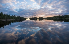Clouds and sunrise reflected in a still lake surrounded by pine trees.